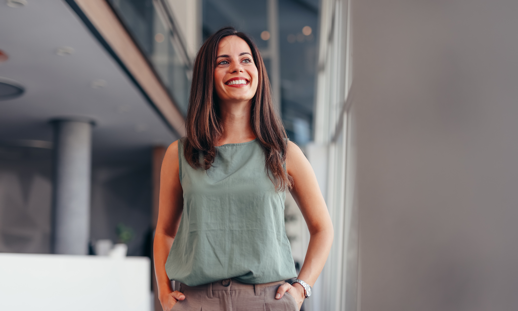 A woman with shoulder-length brown hair, standing with her hands in her pockets and smiling proudly.